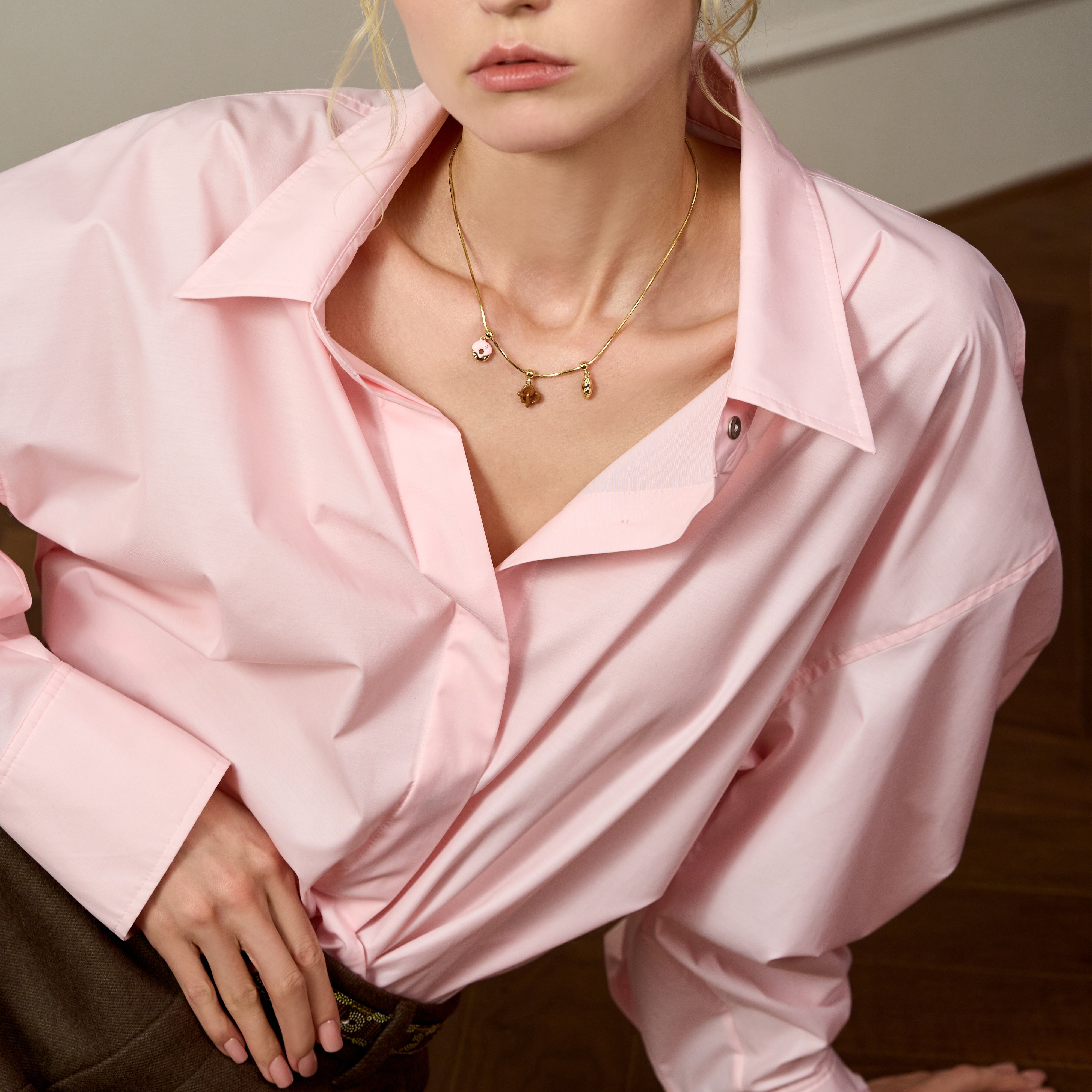 Woman wearing a gold-plated charm necklace with a pink collared shirt and brown pants