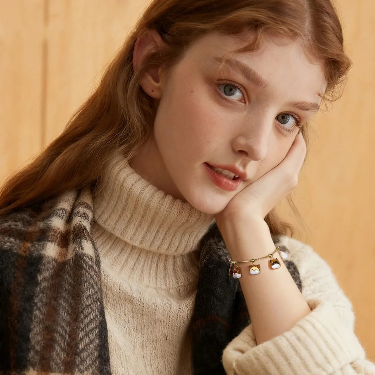 Woman wearing cozy cream turtleneck sweater, plaid scarf, and gold-plated charm bracelet