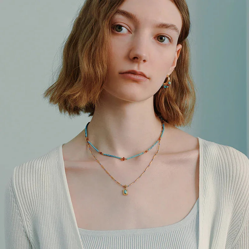 Woman wearing layered gold-plated necklaces with turquoise and bead details, elegant earrings.