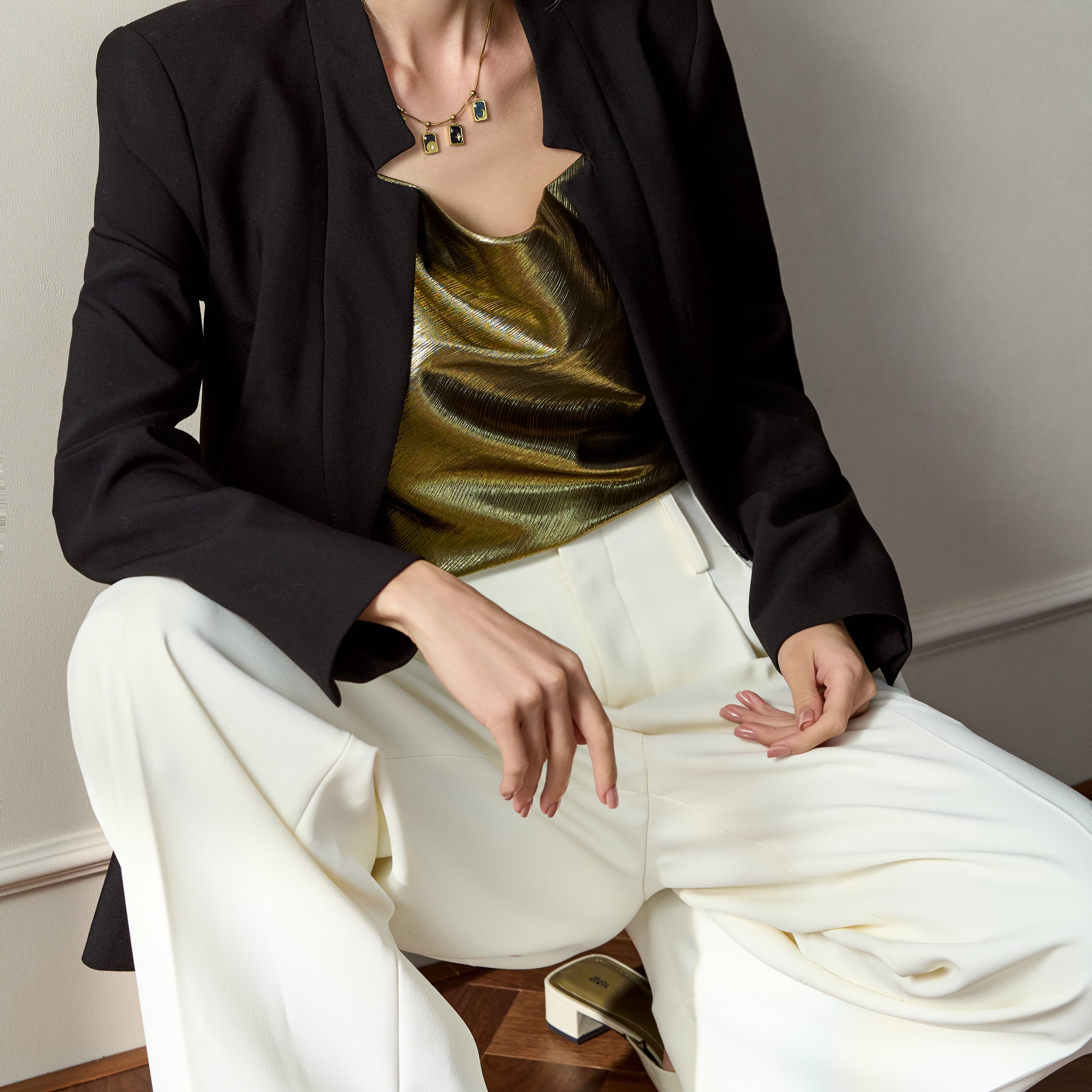 Woman in black blazer, gold metallic top, white trousers, wearing geometric pendant necklace