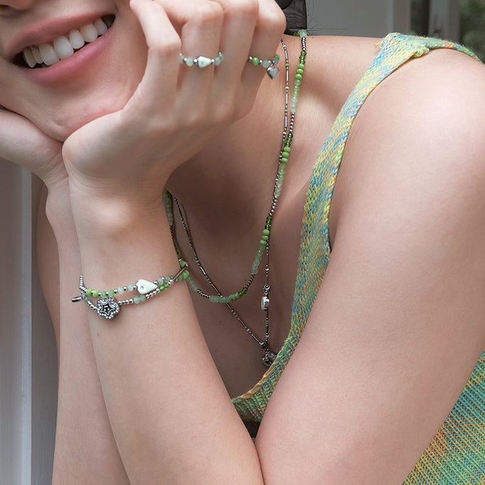 Woman wearing layered green bead necklaces, bracelets, and rings with heart and charm details