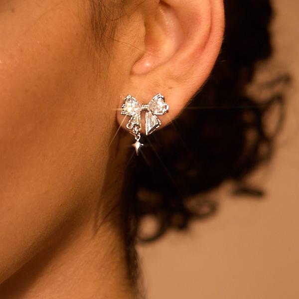 Close-up of a woman's ear wearing a sparkling bow-shaped silver stud earring with a dangling charm.