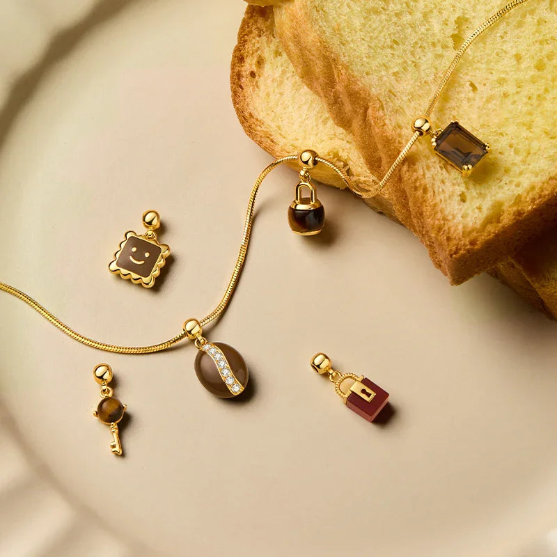 Gold-plated necklaces and cute charm pendants with brown enamel on a beige plate beside bread.