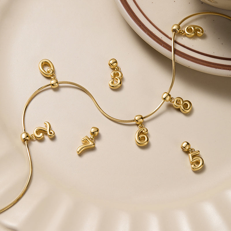 Gold-plated number pendants on a necklace displayed on a cream ceramic plate