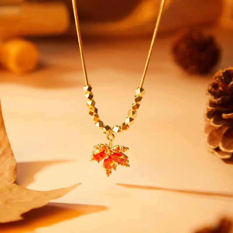 Gold-plated necklace with red crystal maple leaf pendant, styled on a warm autumn background