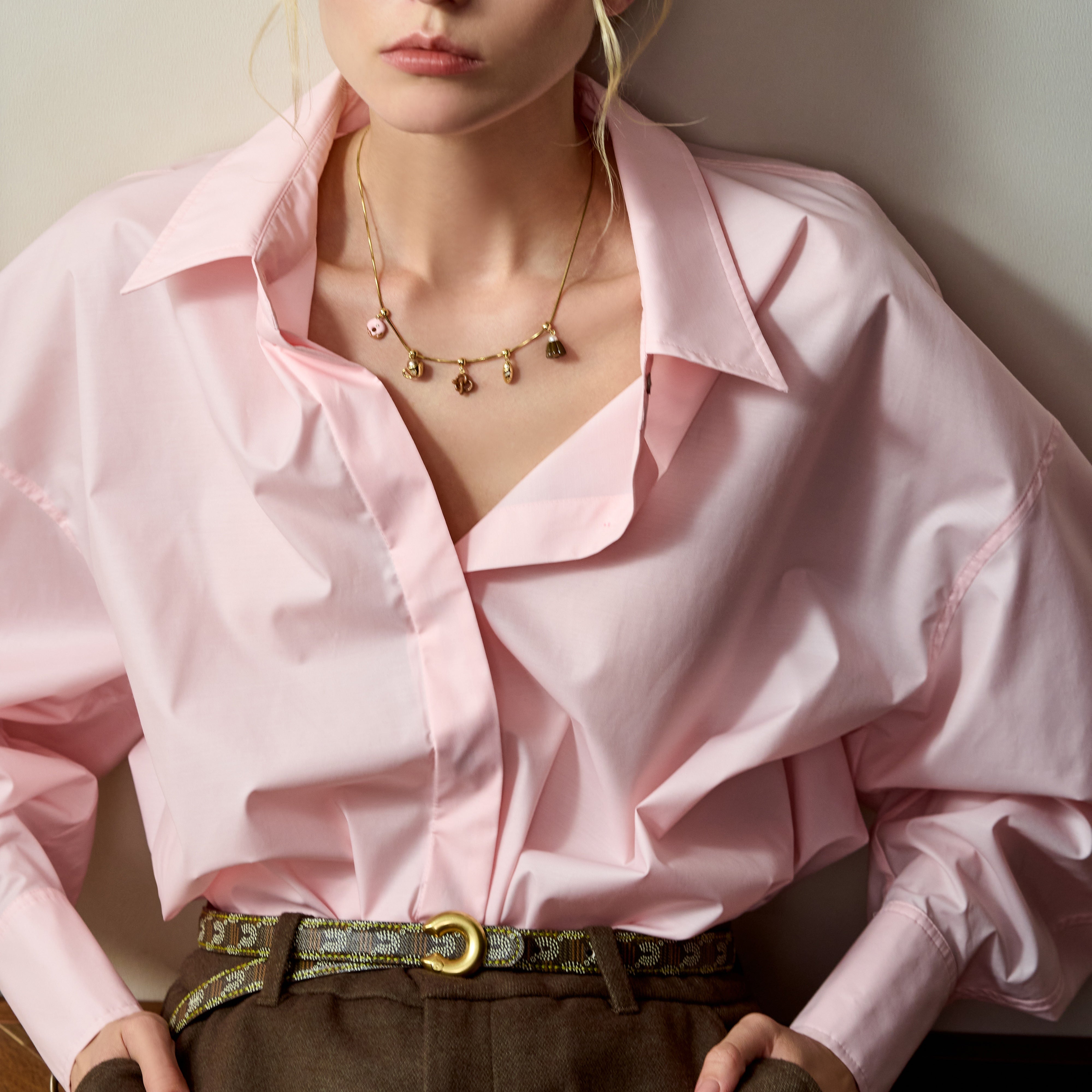 Woman wearing a gold-plated charm necklace, pink blouse, and patterned belt