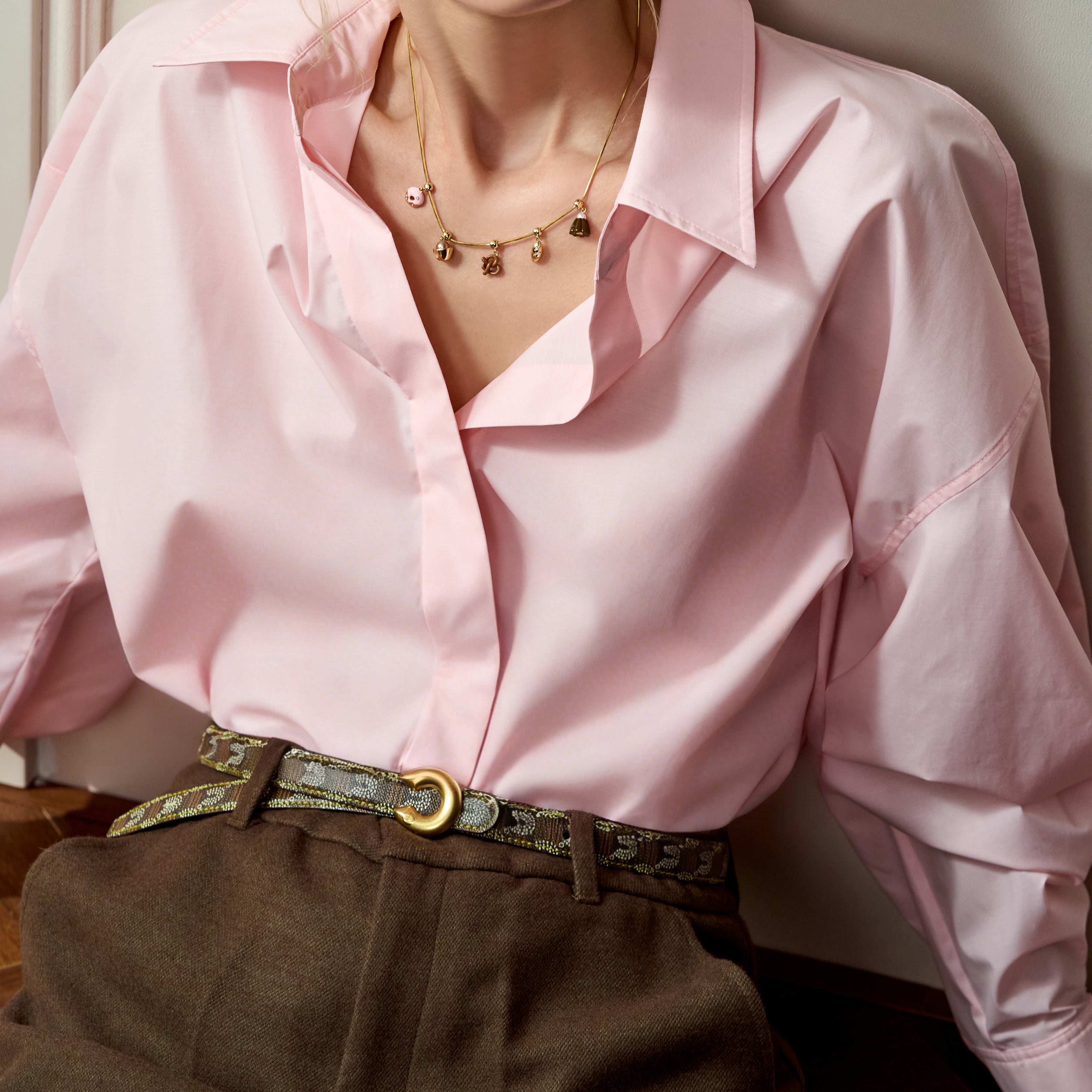 Woman wearing gold-plated charm necklace, pink blouse, and high-waisted brown pants
