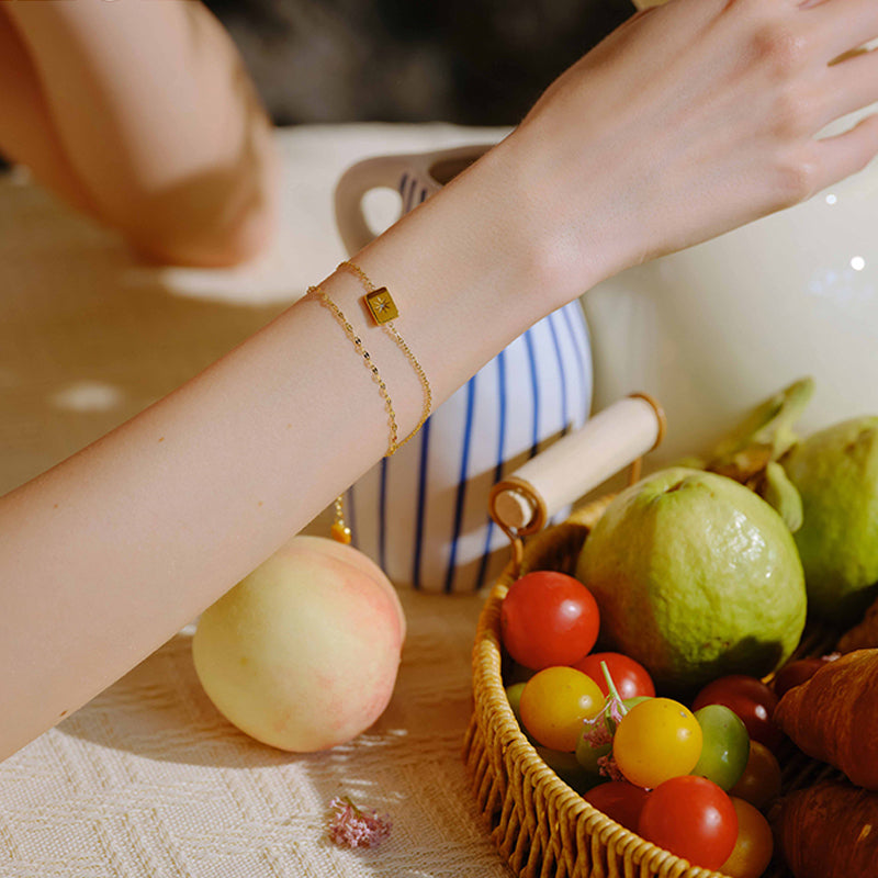 Gold-plated layered bracelet on woman's wrist beside fruit basket and striped teapot
