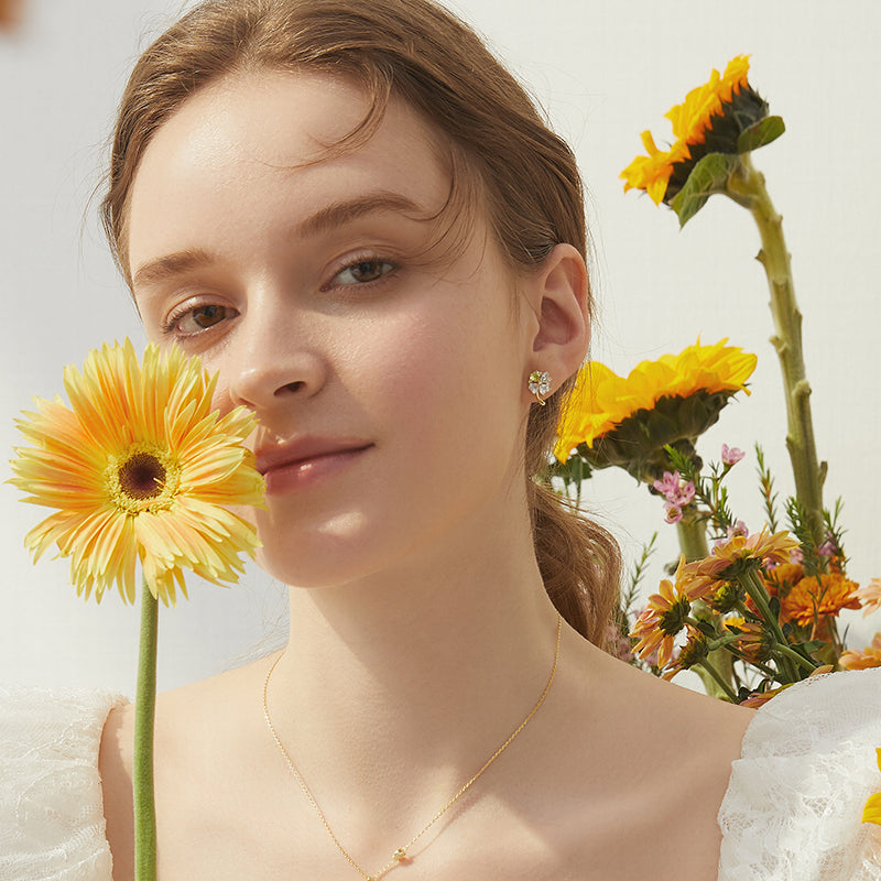 Woman wearing gold-plated necklace and floral earrings, holding a yellow gerbera daisy, with sunflowers in background