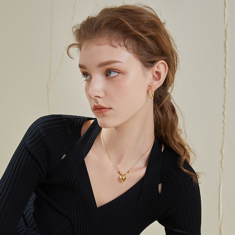 Woman wearing gold-plated hoop earrings and a heart pendant necklace, black top.