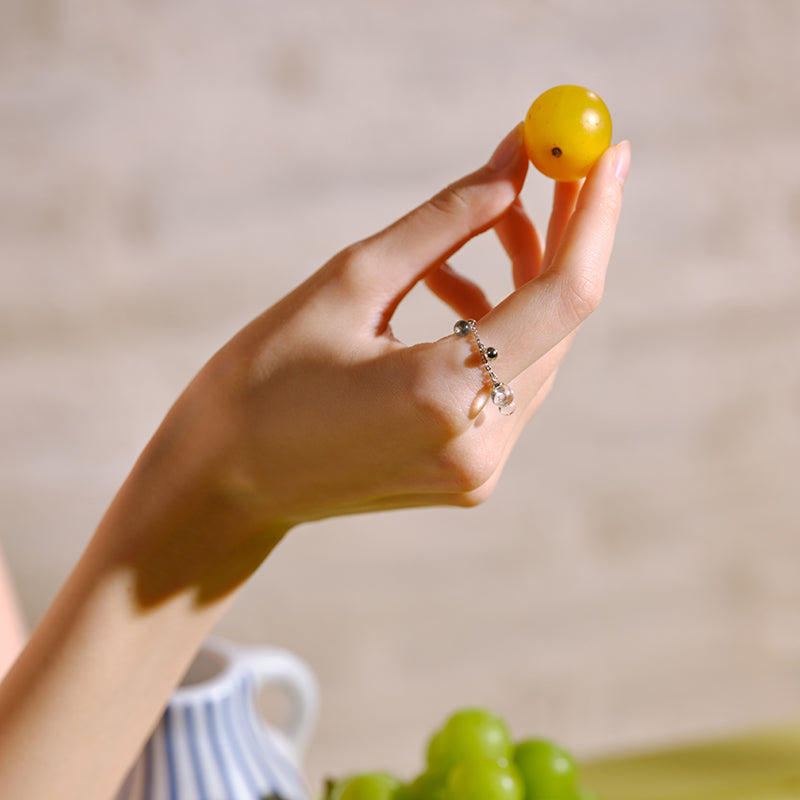 Hand wearing a silver beaded ring with charm, holding a yellow grape, with green grapes in background.