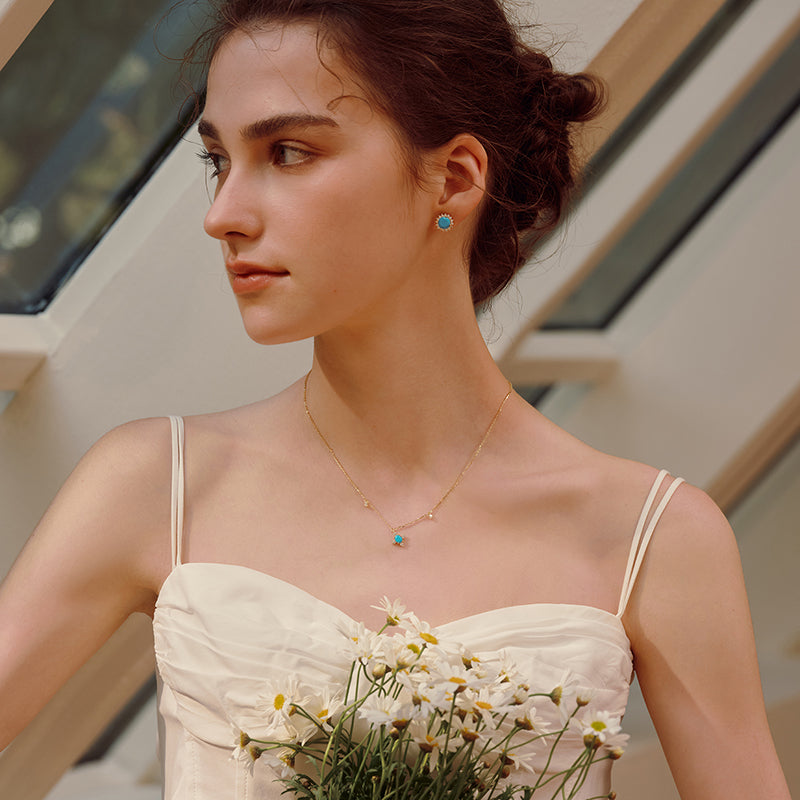 Woman in white dress wearing gold-plated necklace and blue gemstone earrings, holding daisies