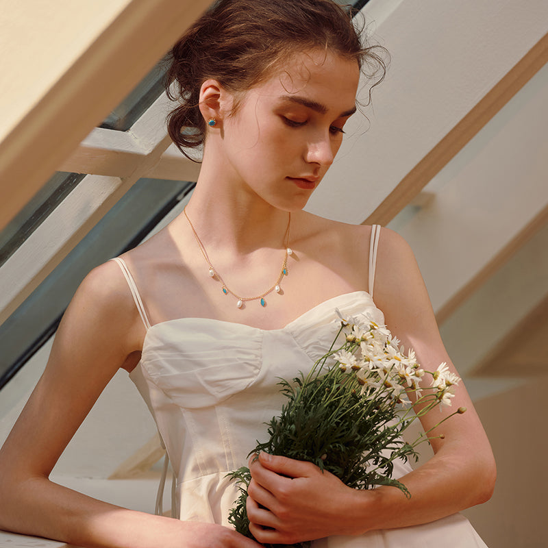 Woman in white dress holding daisies, wearing gold-plated necklace with charms and stud earrings